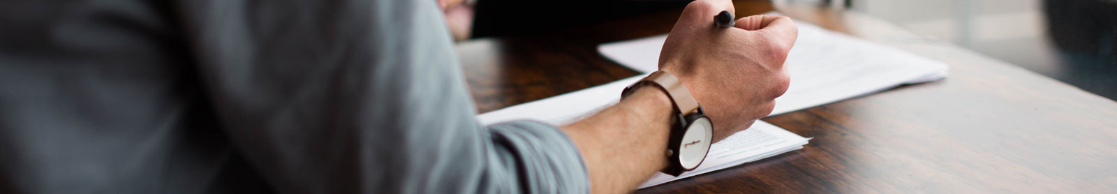 Cropped photo of man's hand holding a pen and wearing wrist watch and sitting at table with papers