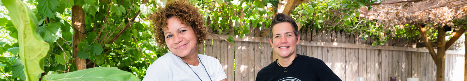 Photo of two women standing outside with lush greenery and a wooden fence behind them