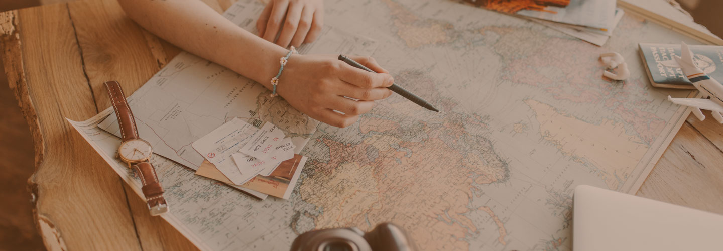 Photo of woman's arm holding a pen and pointing at a large map on a wooden table with travel-related items