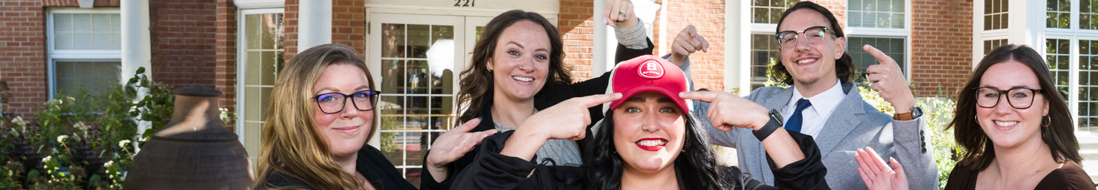 Cropped photo of employees pointing towards their colleague who is wearing a red baseball cap