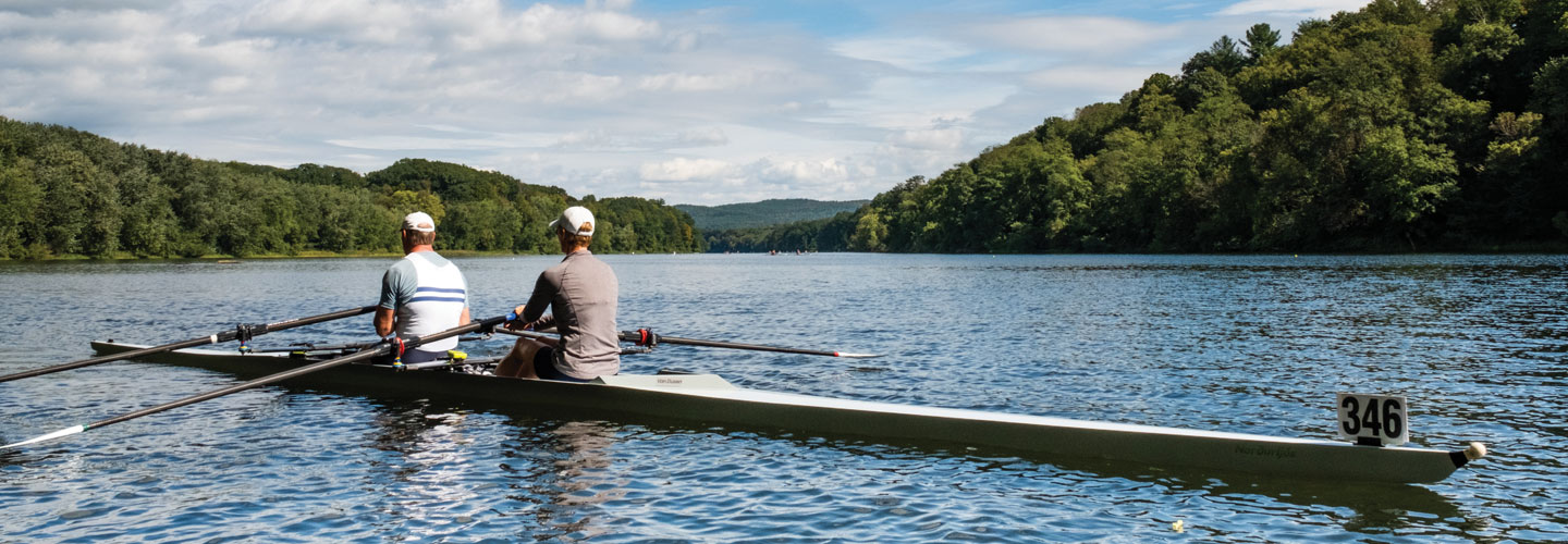 Two men competitively rowing a boat on a lake with a beautiful blue sky and mountains in background