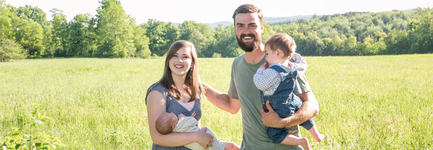 Happy young family with mom holding baby and dad holding toddler. They're standing in a picturesque field with mountains and trees in the background.