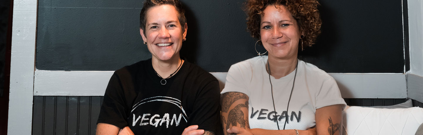Two smiling women standing in front of a blackboard, both women are wearing t-shirts with 'Vegan' on the front