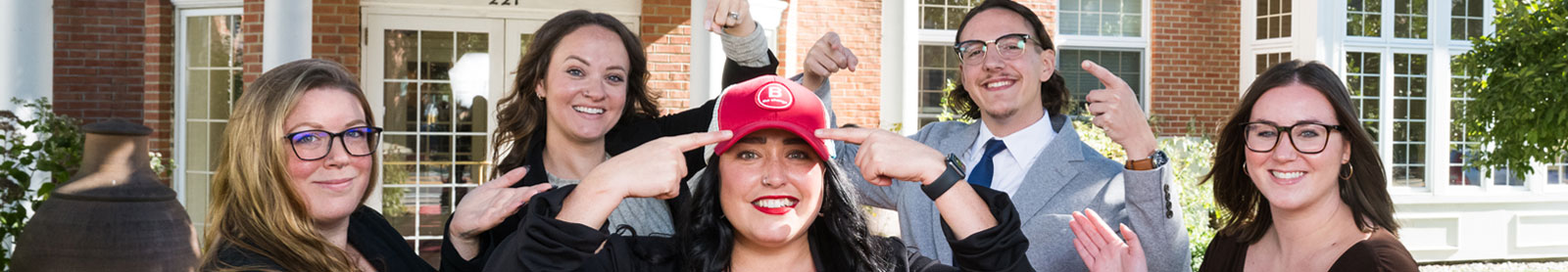 Cropped photo of employees pointing towards their colleague who is wearing a red baseball cap
